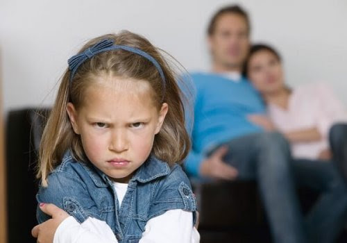 barn uppför sig sämre i sina föräldrars närvaro: flicka tittar argt in i kameran, föräldrar på soffa i bakgrunden