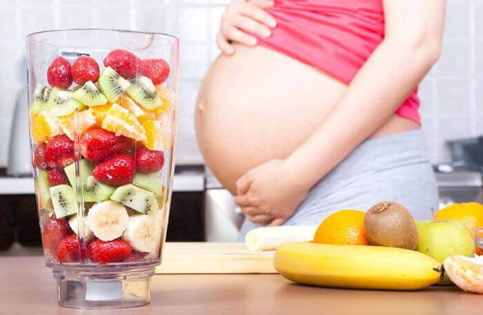 gravid kvinna visar upp magen bredvid en mixer med frukt