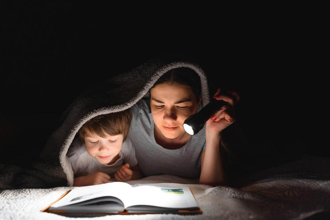 uppmuntra barn att läsa och skriva: mamma och barn läser under filt med ficklampa